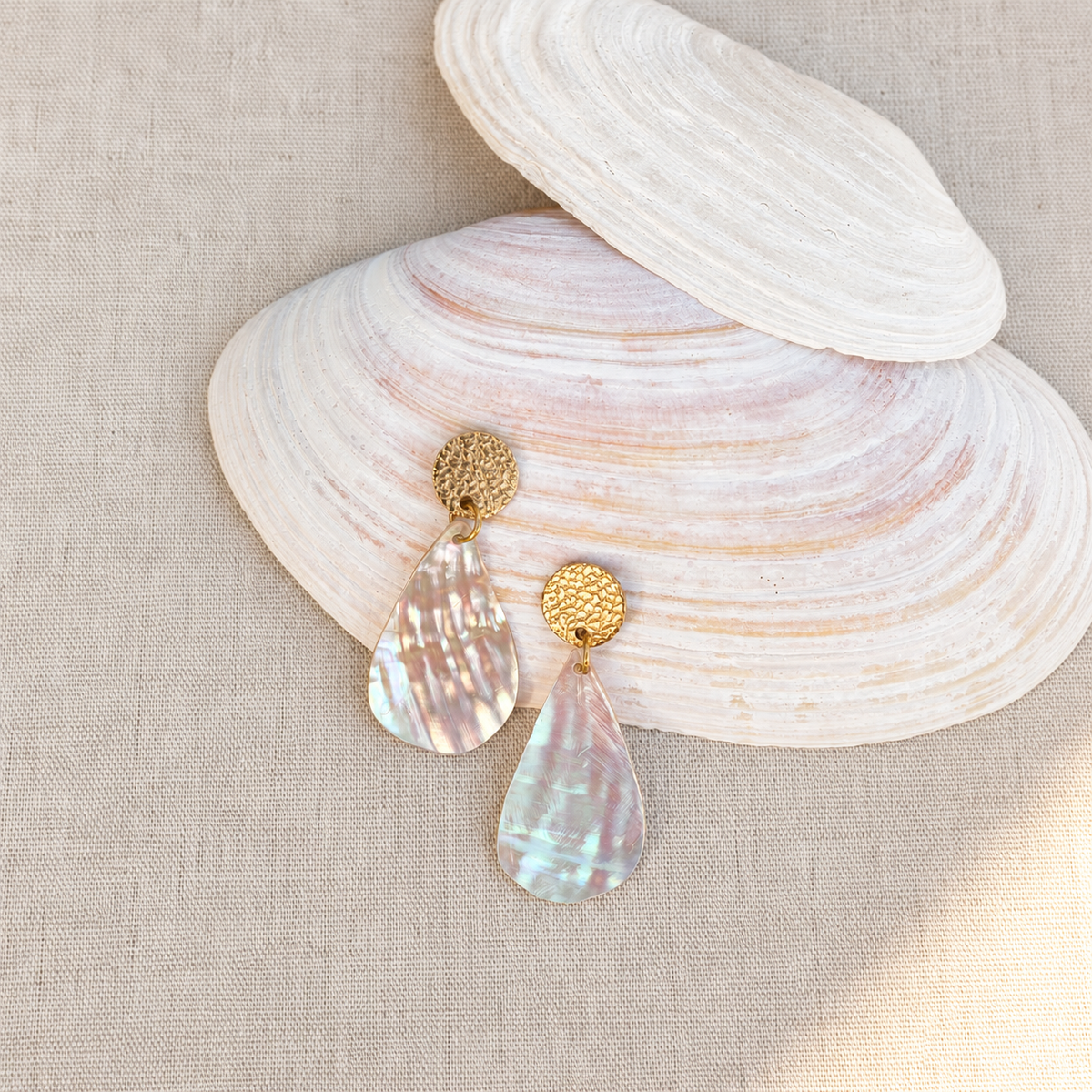 Petites boucles d'oreilles nacre irisée en forme de goutte longue, dorées, posées sur un coquillage. Atelier Sitaël