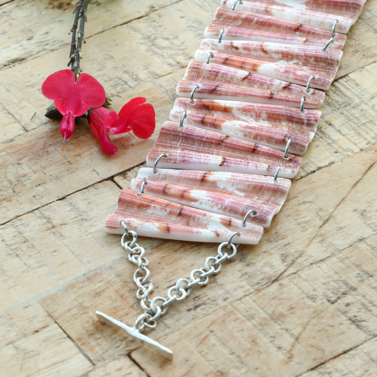 Gros plan d'un bracelet en coquillage rose corail strié et veiné, chaîne et fermoir en T argentés, sur bois brut
