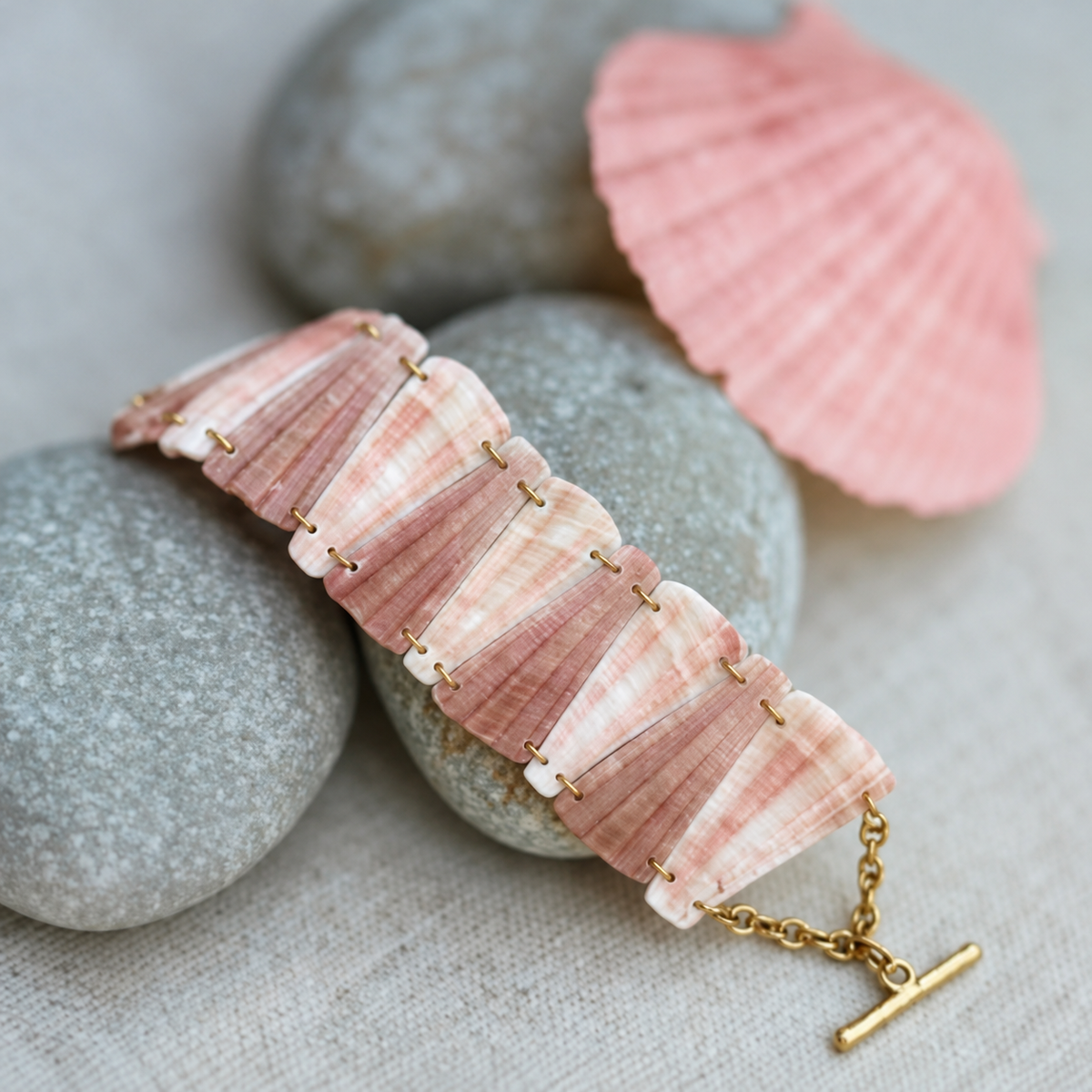 Bracelet en coquillage rose aux maillons trapèzes et fermoir en T doré, posé sur des galets gris à côté d'une coquille rose