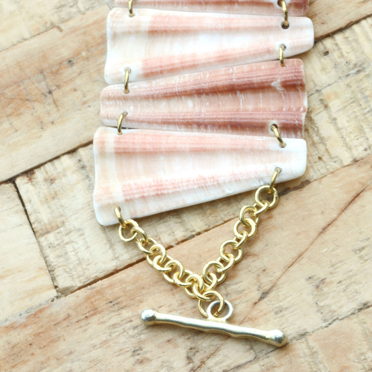 Gros plan d'un bracelet en coquillage rose et blanc strié, maillons trapèzes, chaîne et fermoir en T dorés, sur bois brut