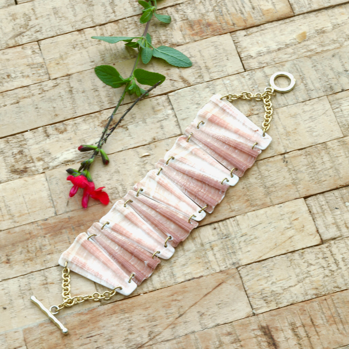 Bracelet entier en coquillage rose et blanc, maillons trapèzes reliés par des anneaux dorés, fermoir en T, posé à plat sur bois brut